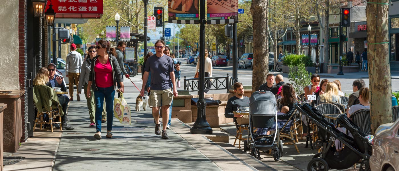 A busy neighborhood retail district with people enjoying sidewalk dining, pushing strollers, and walking dogs.