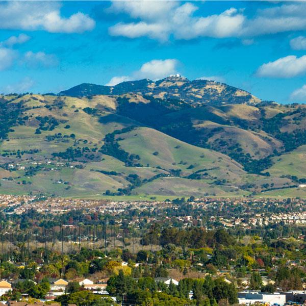 View overlooking Santa Clara Valley with residences in the foreground and green hills in the distance with clouds dotting the blue sky above.